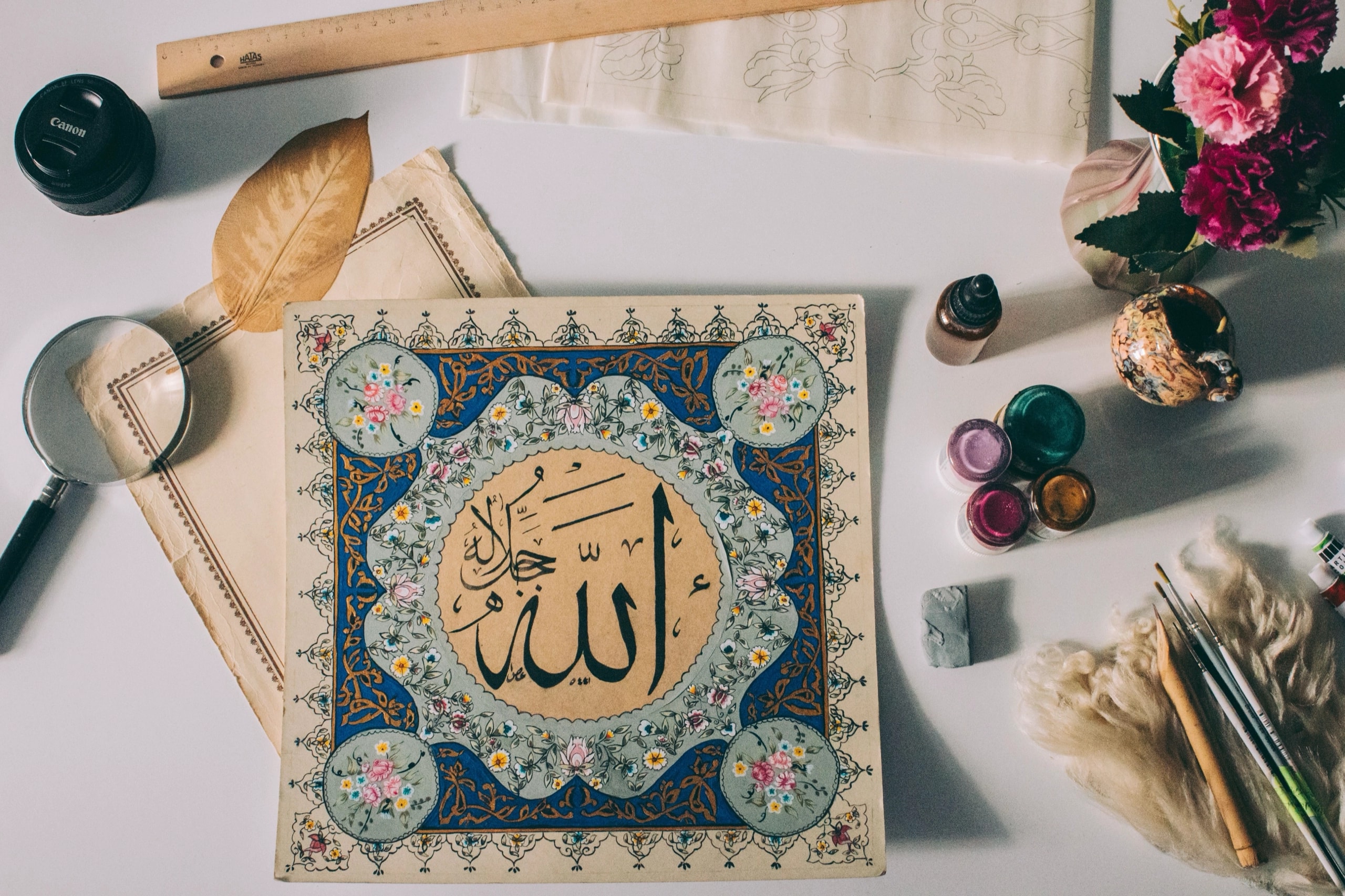 An illuminated Allah calligraphy panel on a desk with brushes, inks and flowers